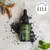 A bottle of SUDTANA’s Bergamot Mint & Turmeric Scalp Revival Treatment in flatlay view with peppermint leaves, turmeric, sesame seeds.