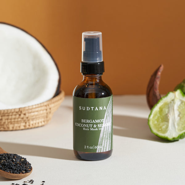 A bottle of Bergamot Coconut & Sesame Hair Mask Oil shown with half a coconut, half a bergamot and a spoonful of sesame seeds for a nourishing hair oil treatment.