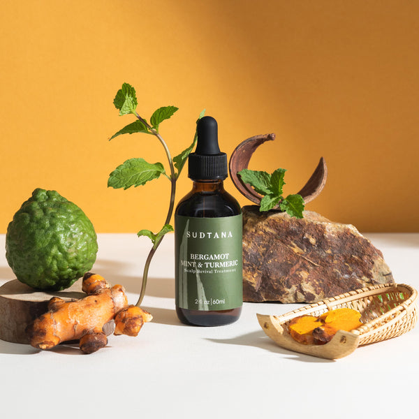 A bottle of SUDTANA's Bergamot Mint & Turmeric Scalp Revival Treatment with peppermint leaves, sliced turmeric, and bergamot for a soothing scalp oil treatment.