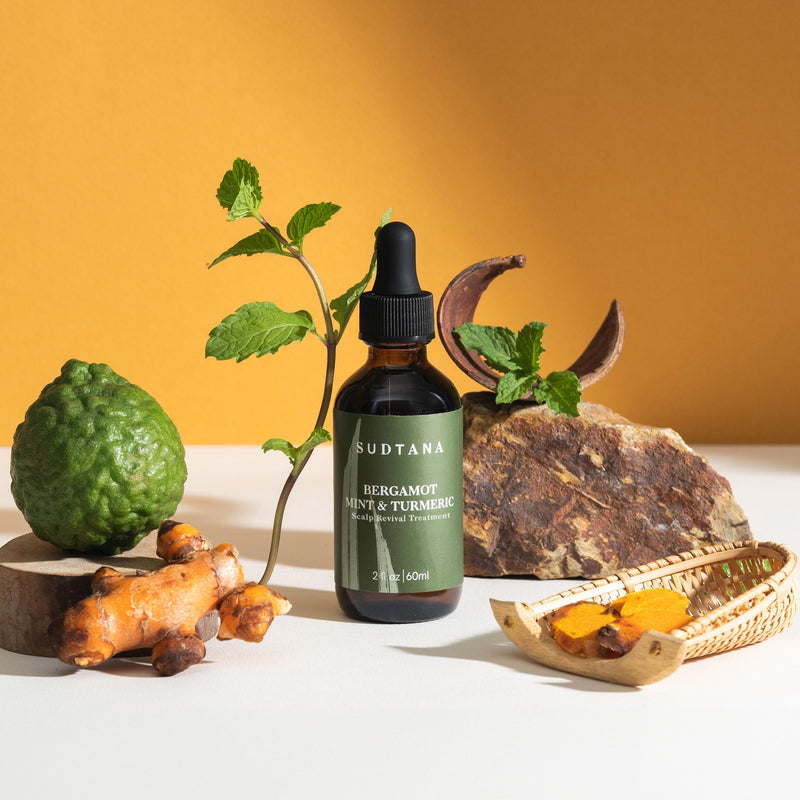A bottle of SUDTANA's Bergamot Mint & Turmeric Scalp Revival Treatment with peppermint leaves, sliced turmeric, and bergamot for a soothing scalp oil treatment.
