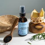 A bottle of Rosemary Ginger & Sesame Uplifting Body Oil displayed with a spoonful of sesame seeds, freshly picked rosemary, and sliced ginger, all sustainably sourced from Thailand.